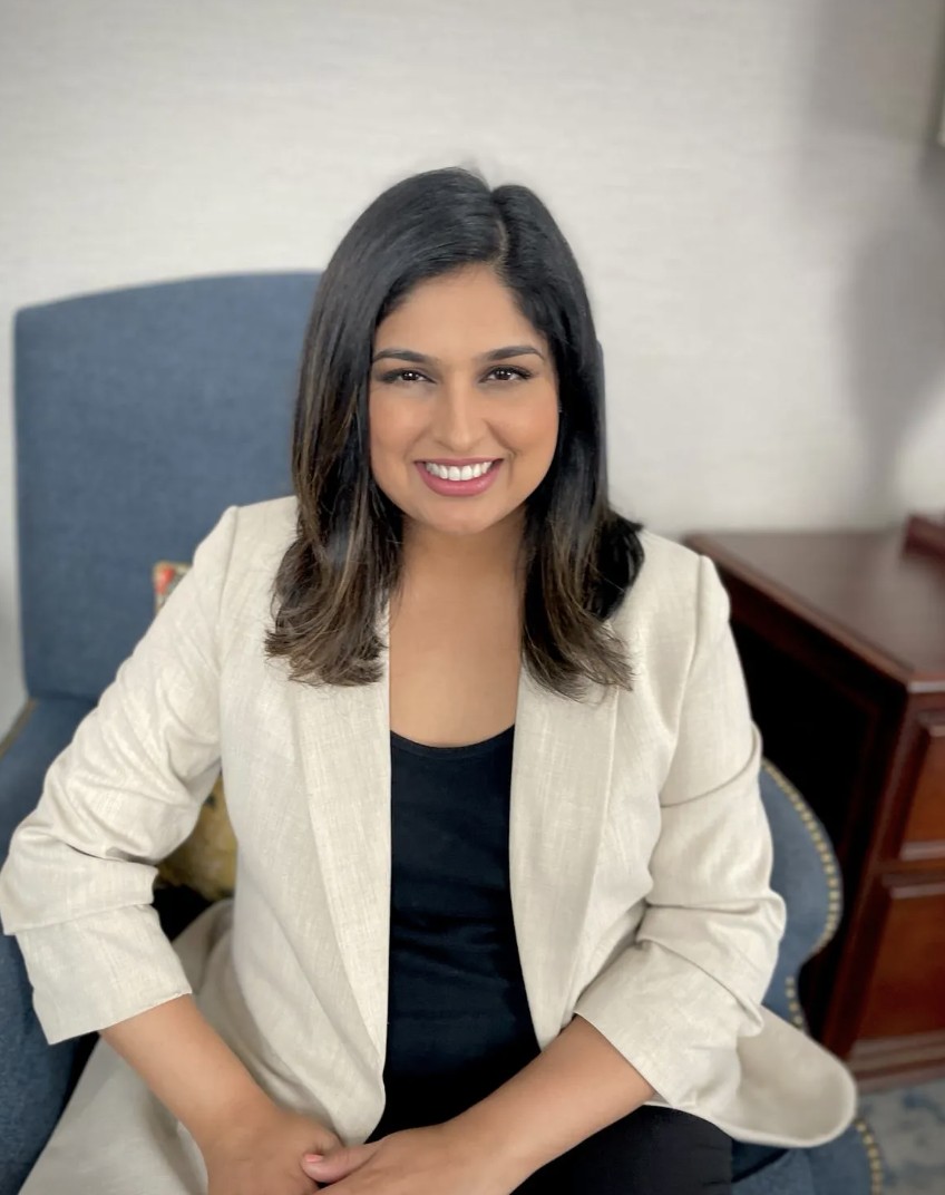 A woman with brown skin and medium-length dark hair wearing a cream blazer over a black shell, smiling and sitting on a medium blue chair