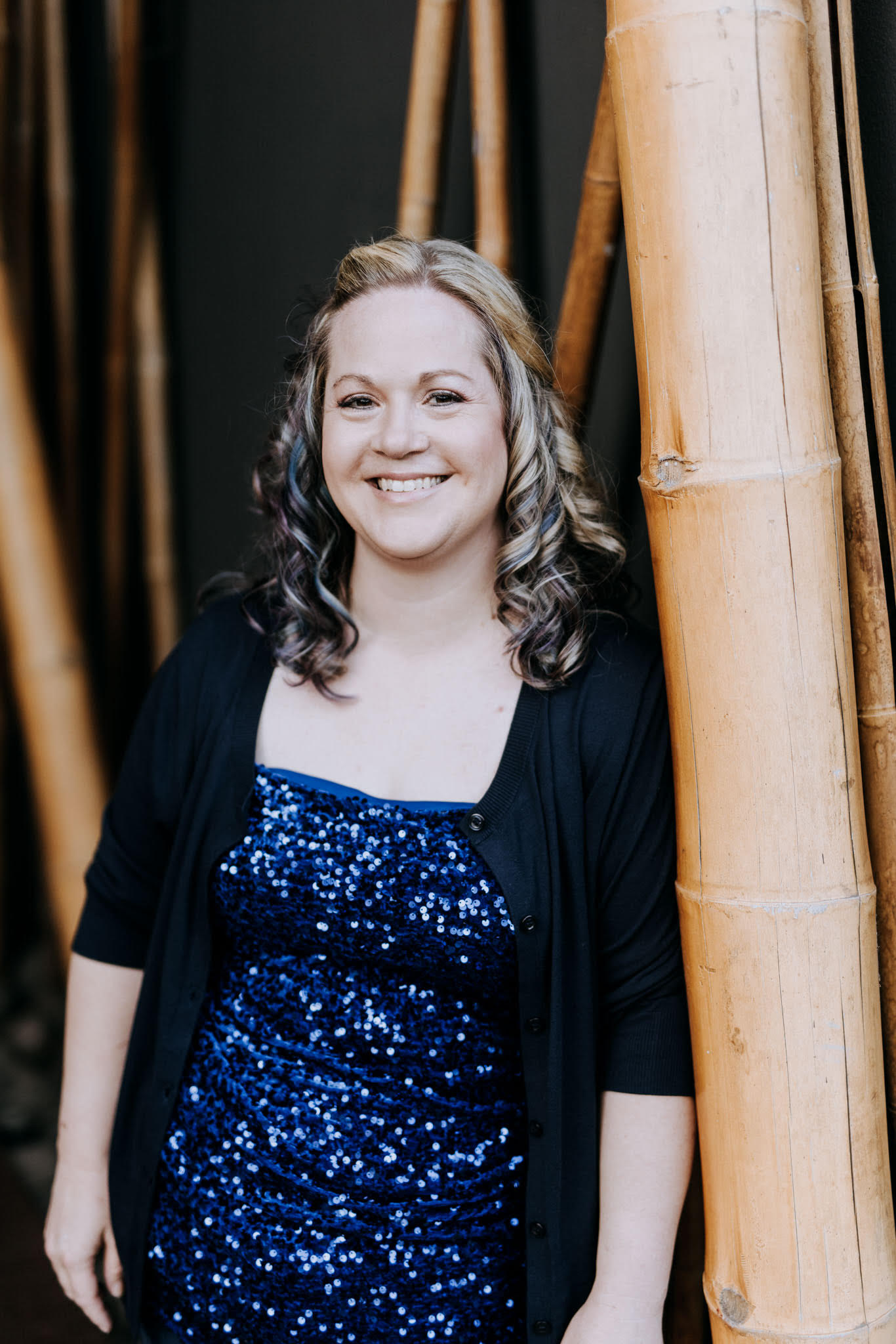 A woman with shoulder-length curly hair and light skin smiling while leaning against bamboo poles, wearing a blue sequined dress and black cardigan.